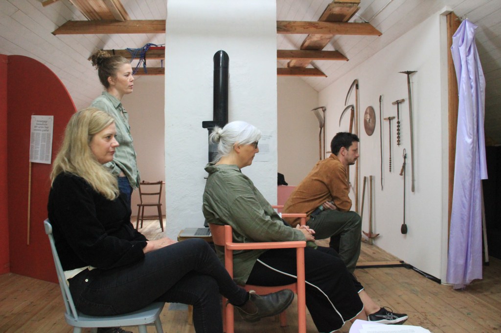 Four visitors sit and stand in the exhibition looking in one direction. Picture is taken from the side. In the background is an installation of tools mounted onto a wall.