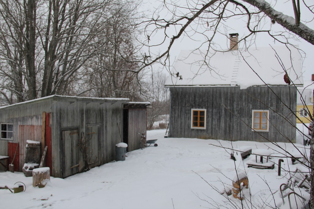The house in Sikås seen from the backside, it has two windows and wooden sides. Left of the house stands a lower storage building made from wood. The ground and roof are covered with snow