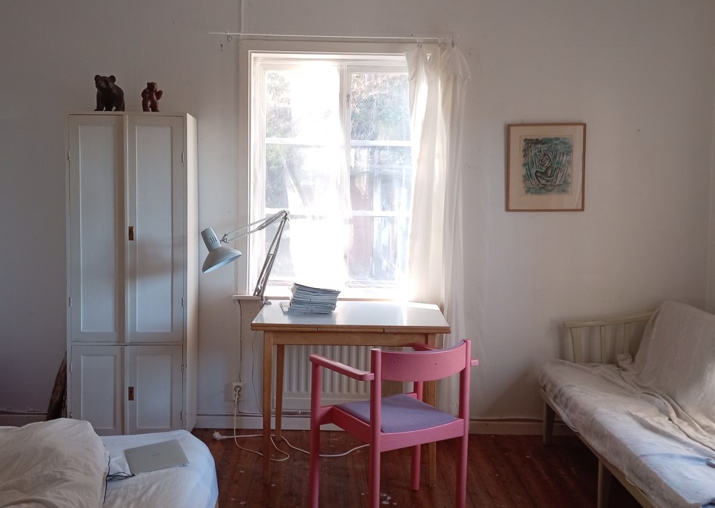 Picture shows a room with a desk, with lamp and chair in front a window. Left of it is a thin white wooden cupboard with two bear sculptures on top of it, on the right is a sofa. In the front a matress and pillow and be seen.