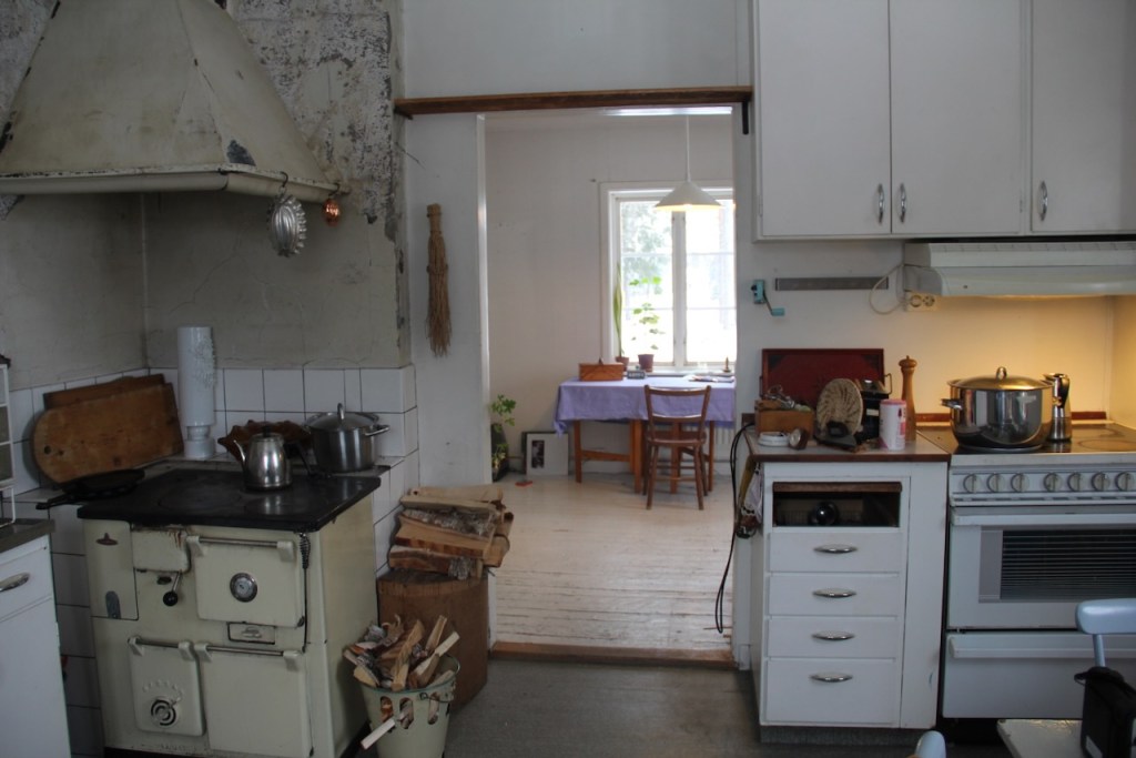 View of the kitchen which has an old wooden stove on the left and kitchen cupboards and an electric stove on the right. Between them an open door offers the view to the next room with a table and chair standing in front of the window.